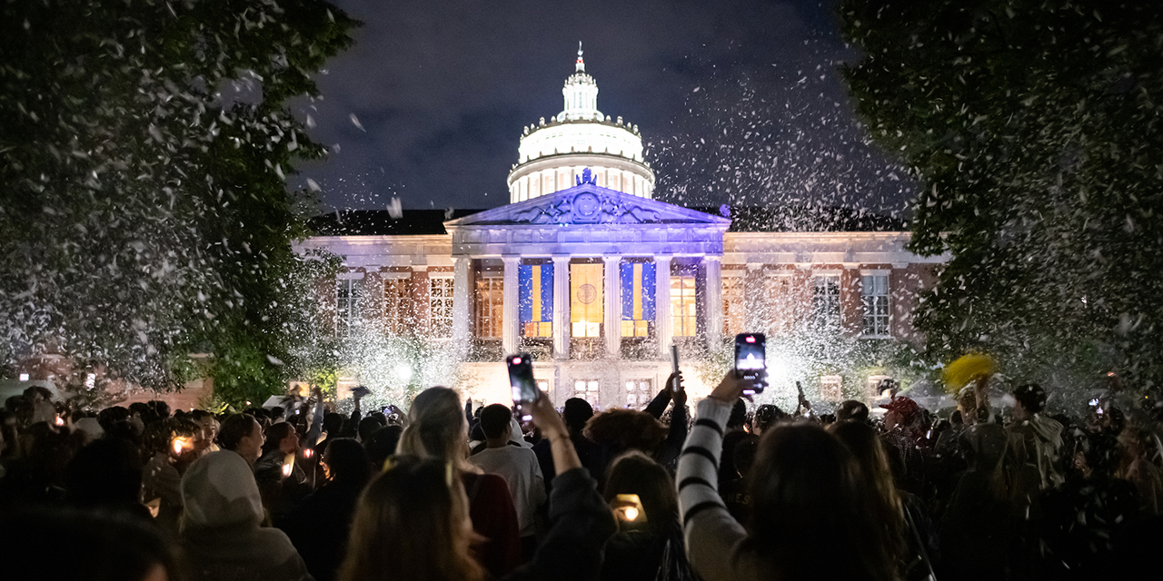 A lit up Rush Rhees Library with students cheering and recording in the foreground as confetti flies through the air