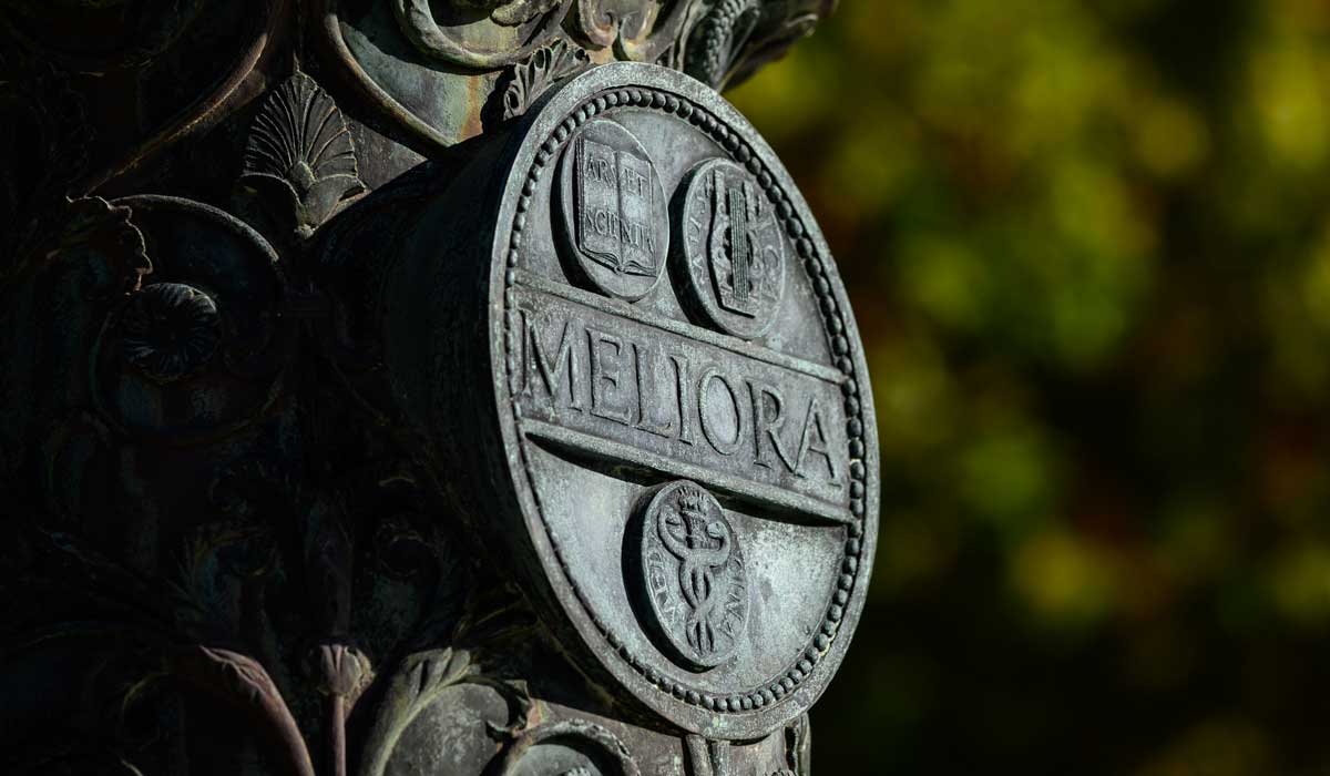 Autumn sunlight catches the weathered bronze of the University of Rochester seal, illuminating Meliora—ever better—against a soft backdrop of golden-green leaves.