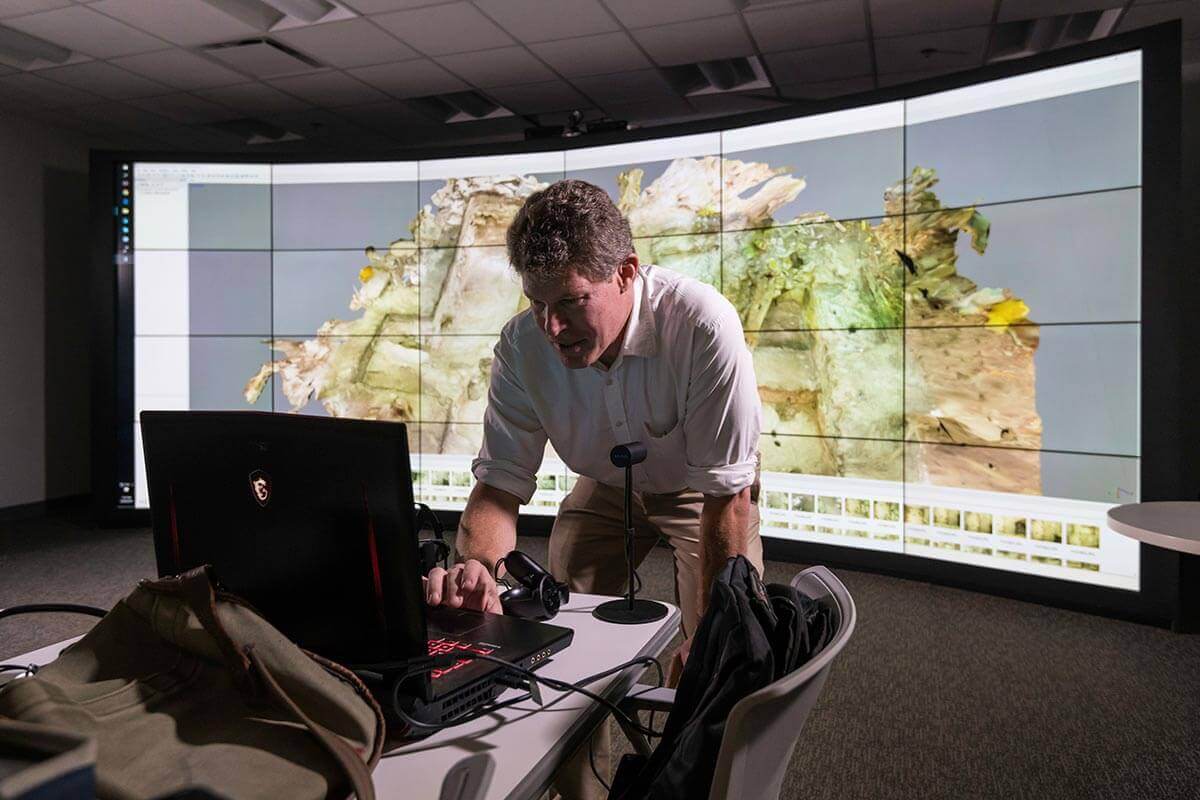 Man leaning over laptop with curved wall monitor display in background at University of Rochester