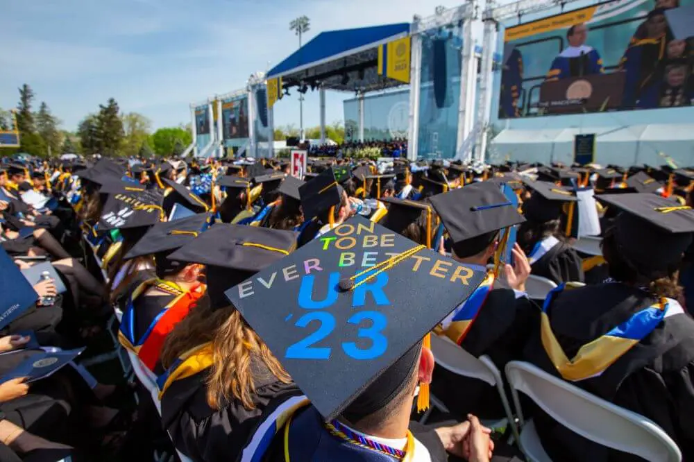 Class of 2023 graduates looking on towards the stage. In the foreground, a graduation cap that says On to Be Ever Better UR '23.