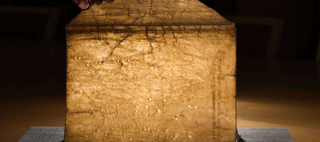 Hand holds up page of medieval manuscript from the Richard Kaeuper Collection as light filters through.