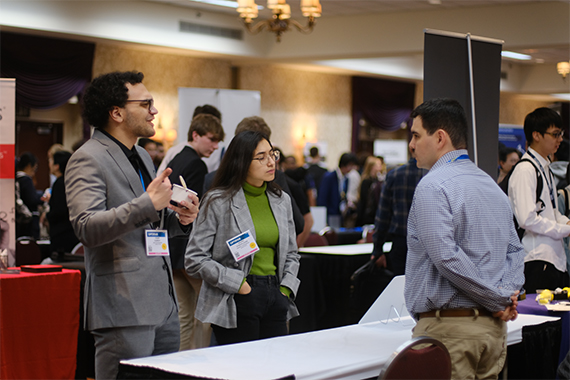Two PhD students speak to an industry representative in a crowded ballroom