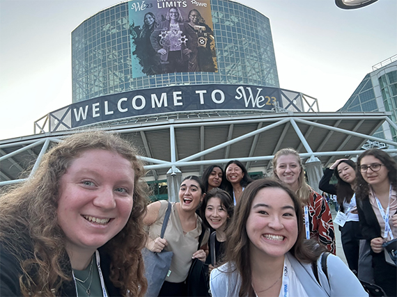 Nine Rochester students pose for a selfie in front of a convention center with a banner that says "WELCOME TO We23"