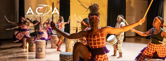 dancers in colorful dress hold drumsticks over drums.