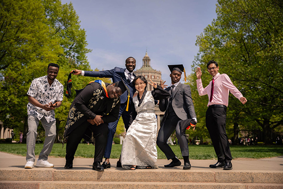 A group of six international gradautes, dressed in caps, gowns, and dress clothes, joyfully pose on a sunny day in front of the Eastman Quad.