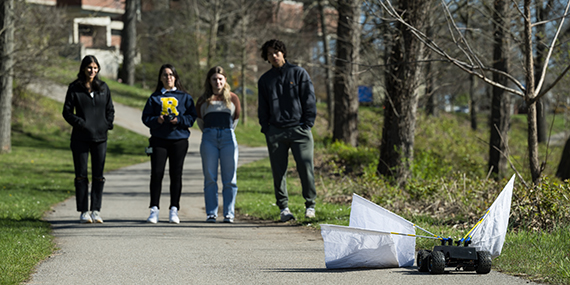 A radio controlled car with white flannel flags attached and four students in the background controlling it on a bike path.