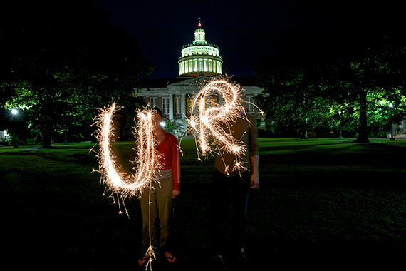 At night, two people use sparklers to draw the letters U and R while standing in front of an illuminated Rush Rhees Library.