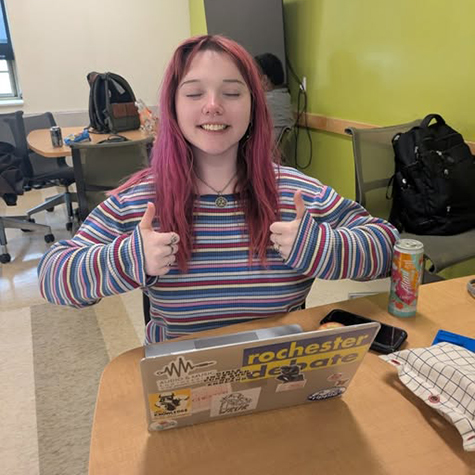 A person smiles at the camera from behind a laptop while sitting at a table.