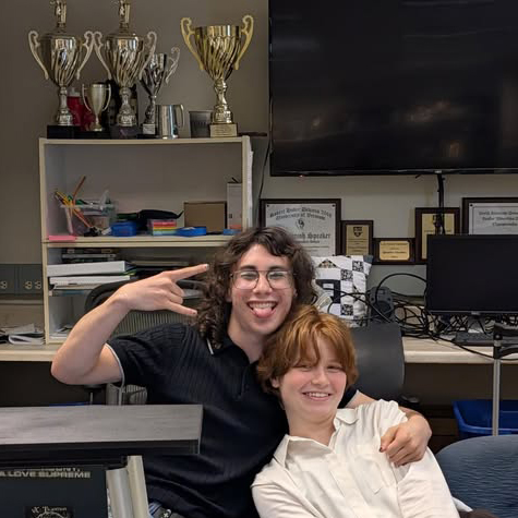 One person has their arm around another while posing for the camera with a desk, TV, and trophy in the background.