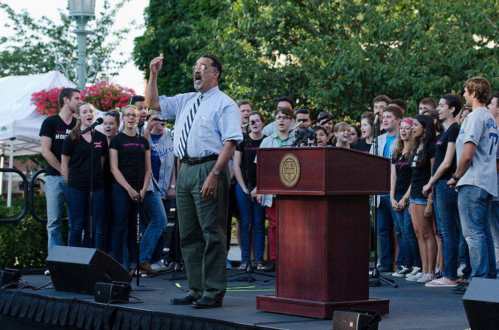 Paul BUrgett on stage outside, leading student singing groups