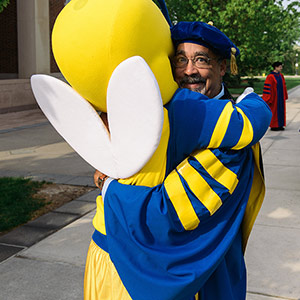 Paul Burgett in commencement robes is hugged by Rocky yellowjacket mascot