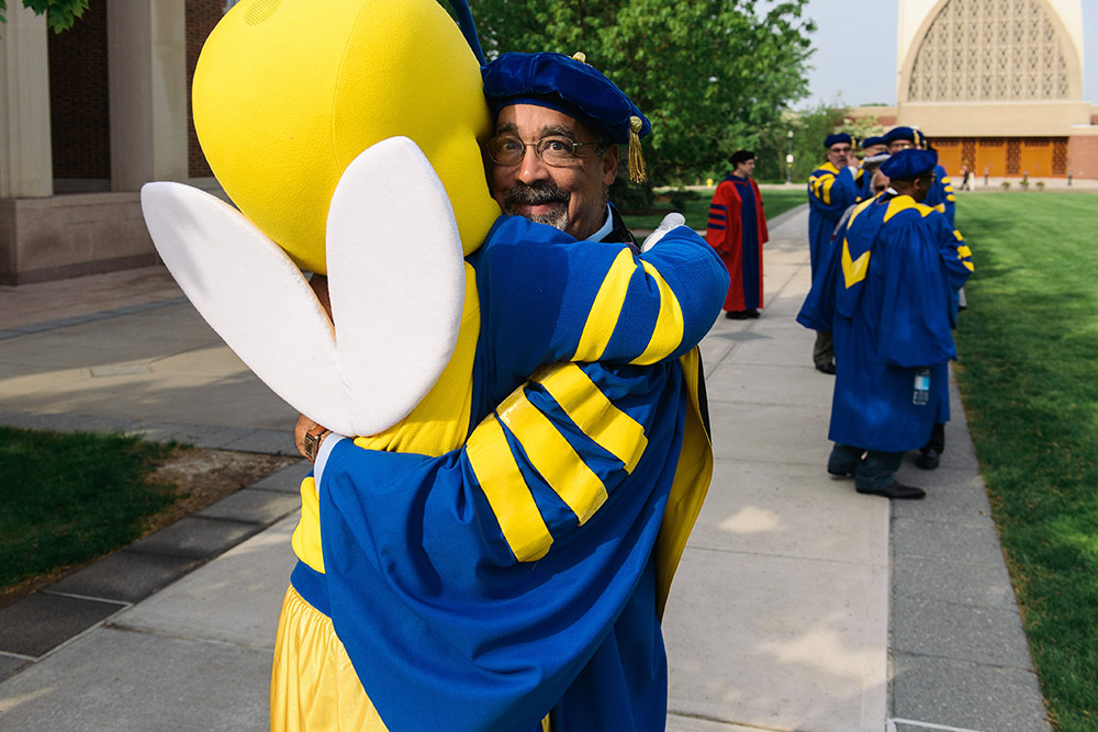 Paul BUrgett in graduation robes is hugged by the Rocky yellowjacket mascot