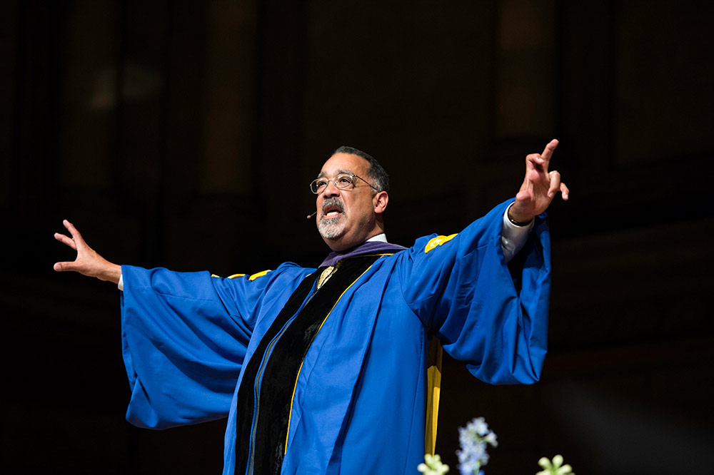 Paul BUrgett in graduation robes gestures while giving a speech