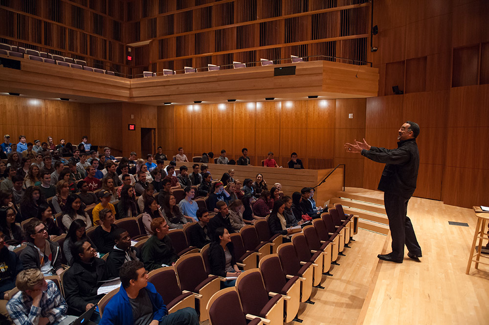 Paul Burgett gestures while giving a speech on stage in front of a large group of students