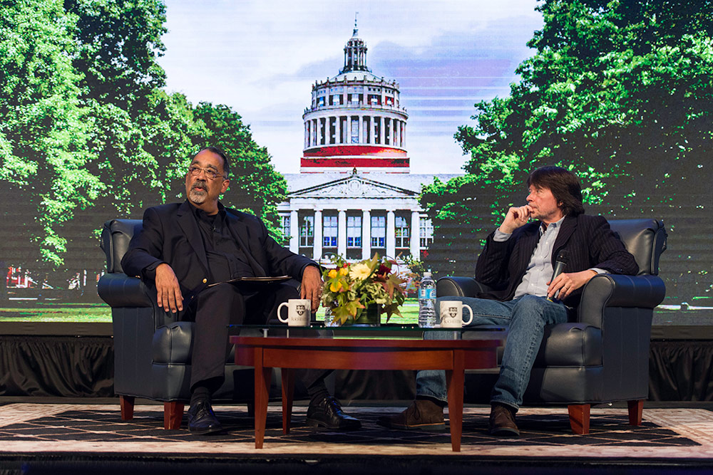 Paul Burgett seating in an armchair on stage with Ken Burns