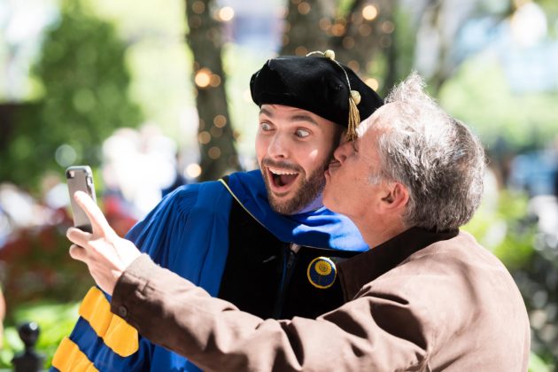 man in graduation garb gets a kiss whil taking a selfie