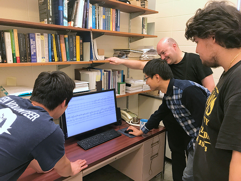students and professor standing around laptop with musical notes on the screen