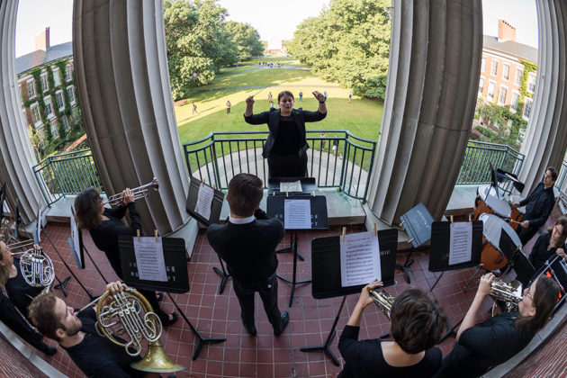 conductor raises arms and faces musicians playing on a balcony