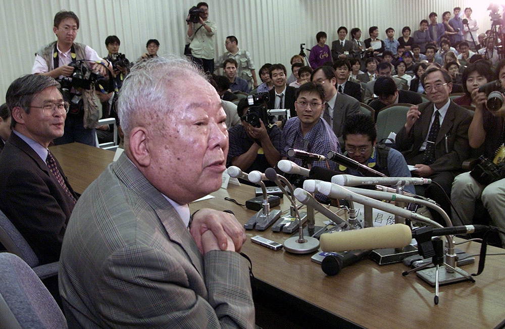 man at a table in front of mics and reporters