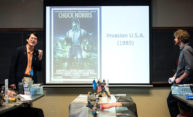 two instructors stand in front of a class with a screen showing a poster for the Chuck NOrris movie INVASION USA