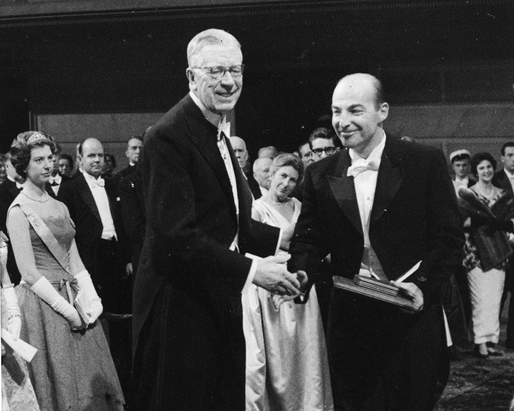 black and white photo of otwo men shaking hands in front of formally dressed audience
