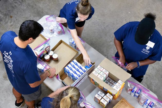 photo looks down from above at four people in matching University of Rochester r-shirts packing boxes with food