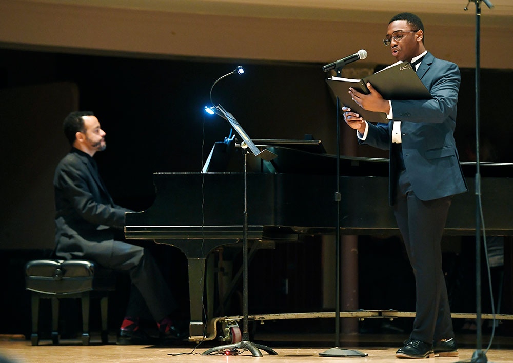 two musicians on stage, one playing the piano the other singing and holding a book of music