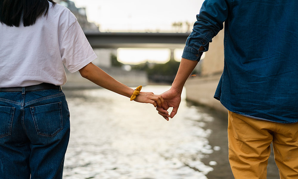 a couple holding hands loosely while walking along a waterfront.