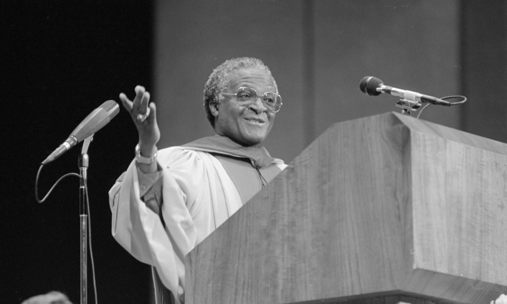 Black-and-white archival photo of Desmond Tutu in University of Rochester commencement robes and at a podium.