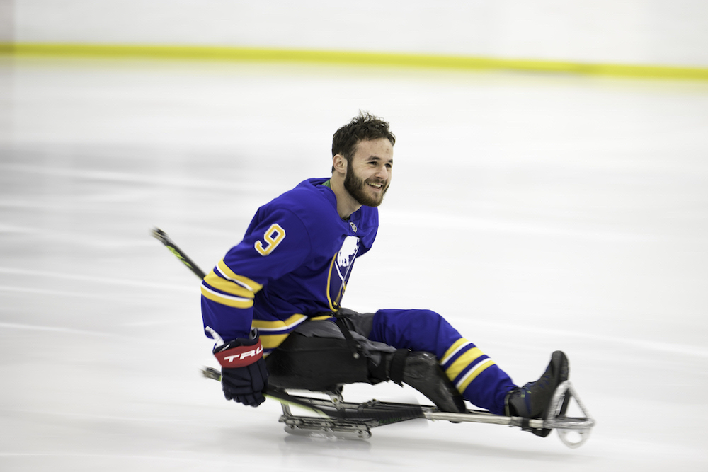 Para ice hockey athlete on skates on the ice, with a hockey stick. Wearing a blue and gold jersey