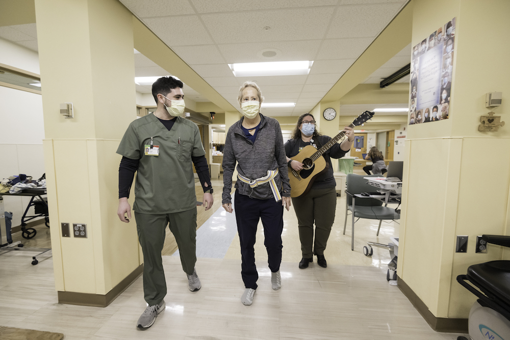 Three people in masks and scrubs walking down a hospital hallway. The woman on the right is carrying a guitar.