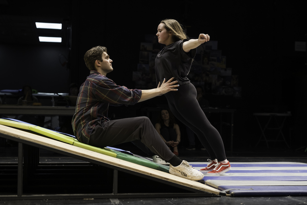 Male college student sits upright on incline on a stage, balancing female college student who has her arms spread. Teacher looks on from background