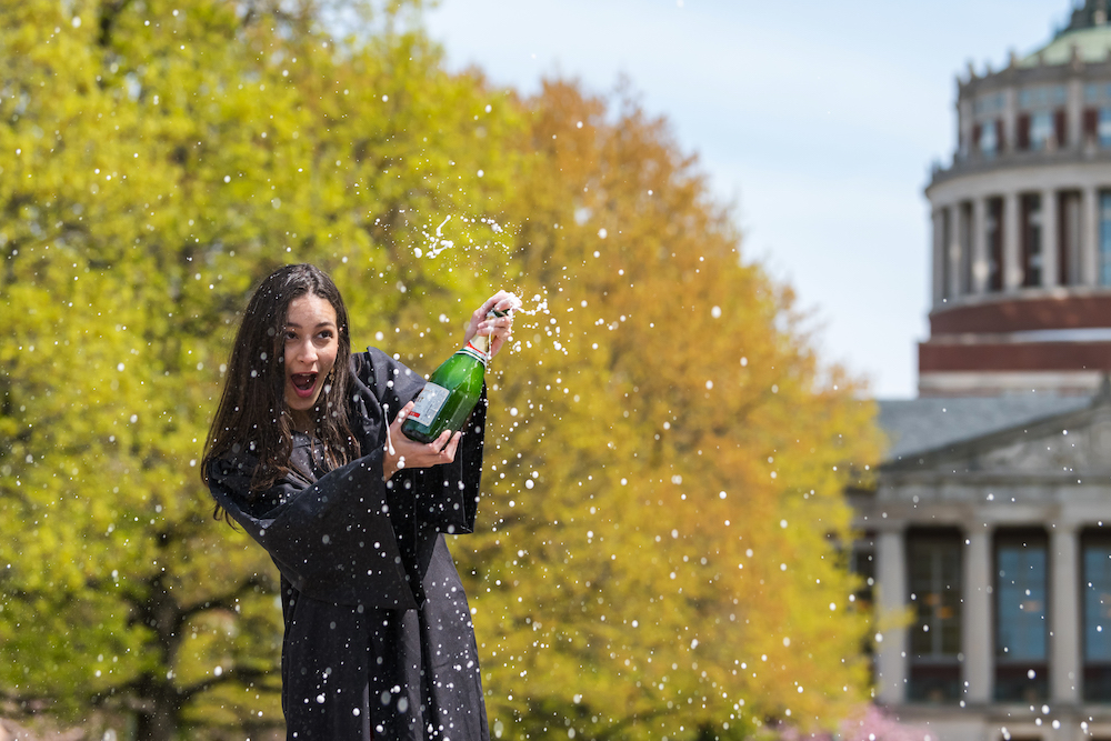 Female student in black graduation gown pops a bottle of champagne