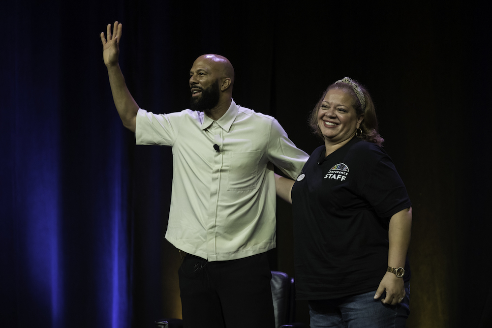 Man and woman stand on stage, arms around each other, waving to audience