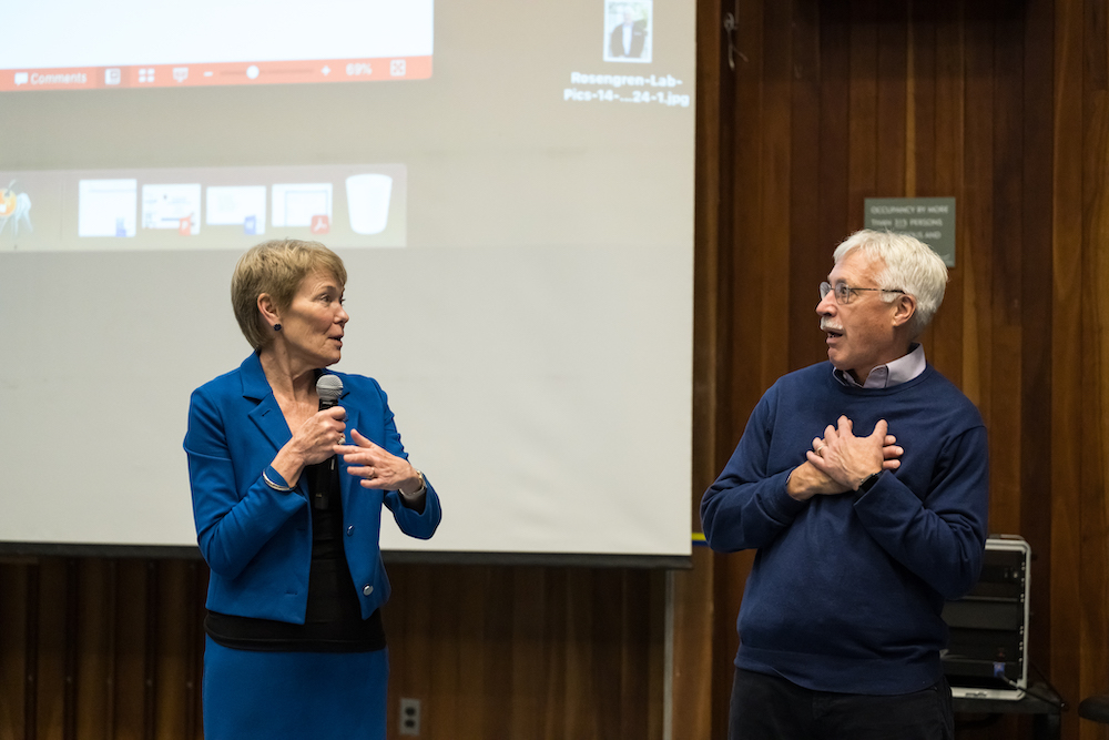 Woman in blue suit stands with microphone next to a man in a blue sweater feigns surprise