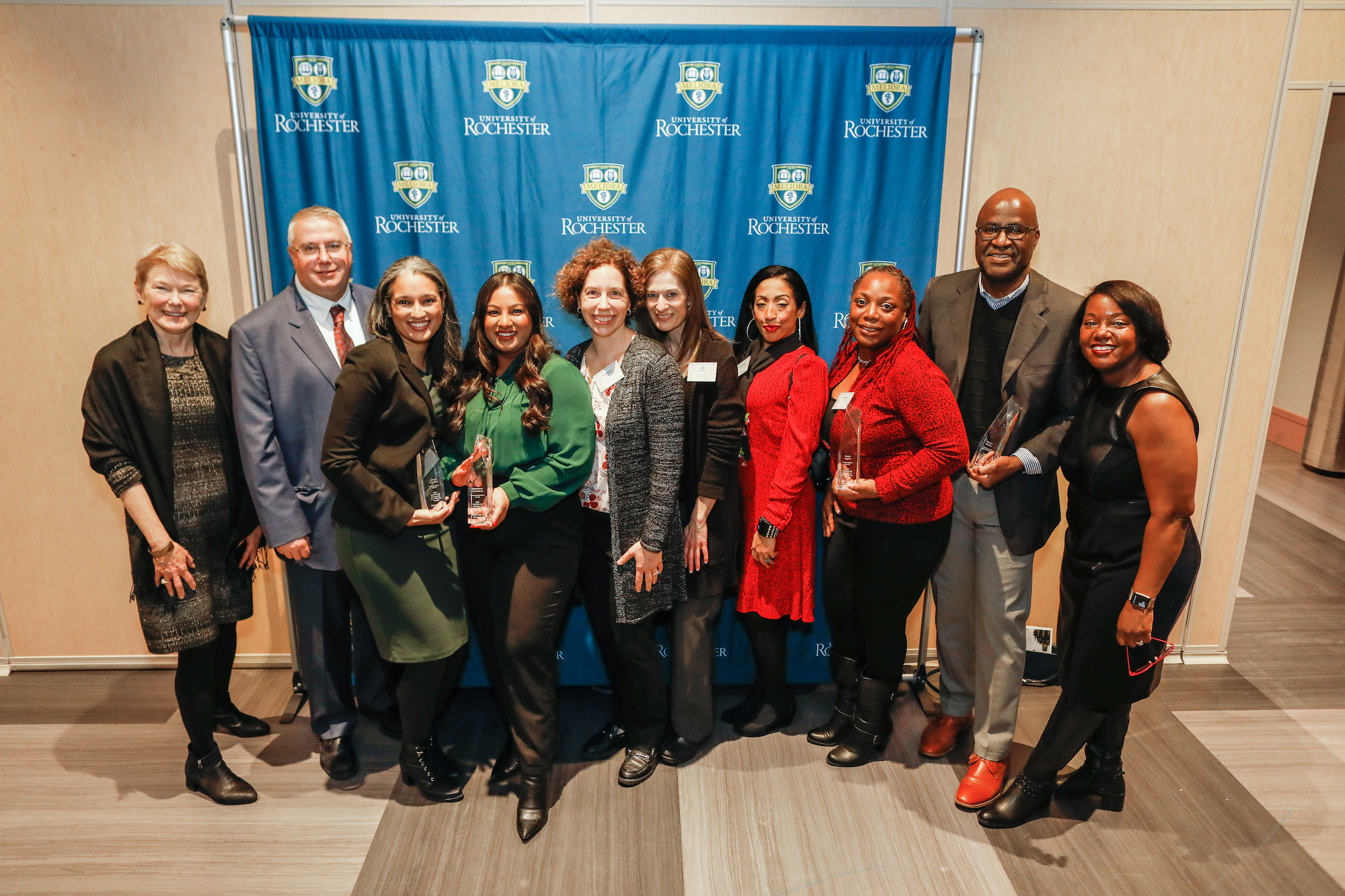 Professionally dressed men and women hold glass awards in front of a blue and gold backdrop