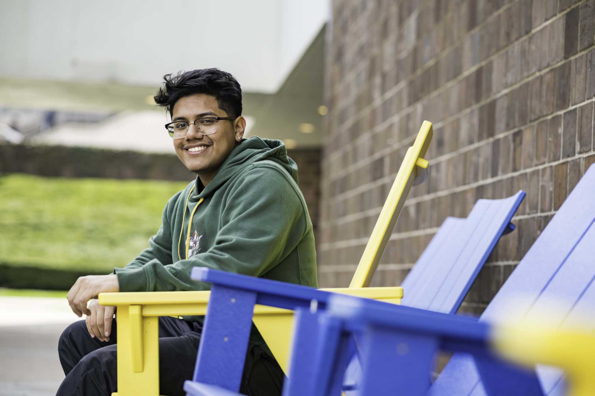 First-generation student Justin Pimentel seated outdoors in a yellow Adirondack chair and smiling at the camera.