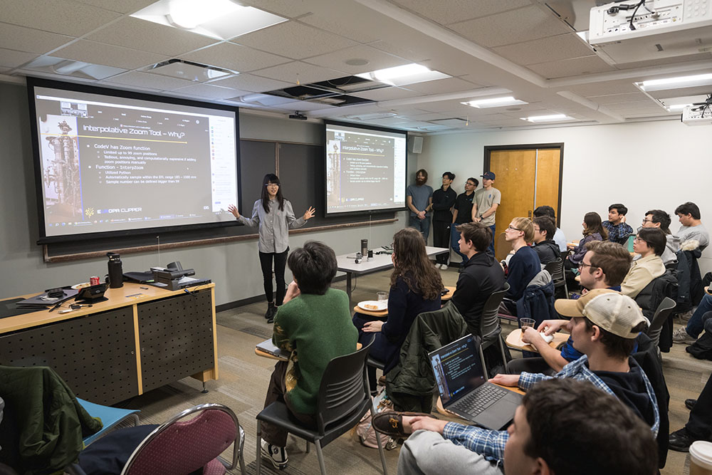 Female PhD student stands before classmates, giving presentation of her group's lens design.