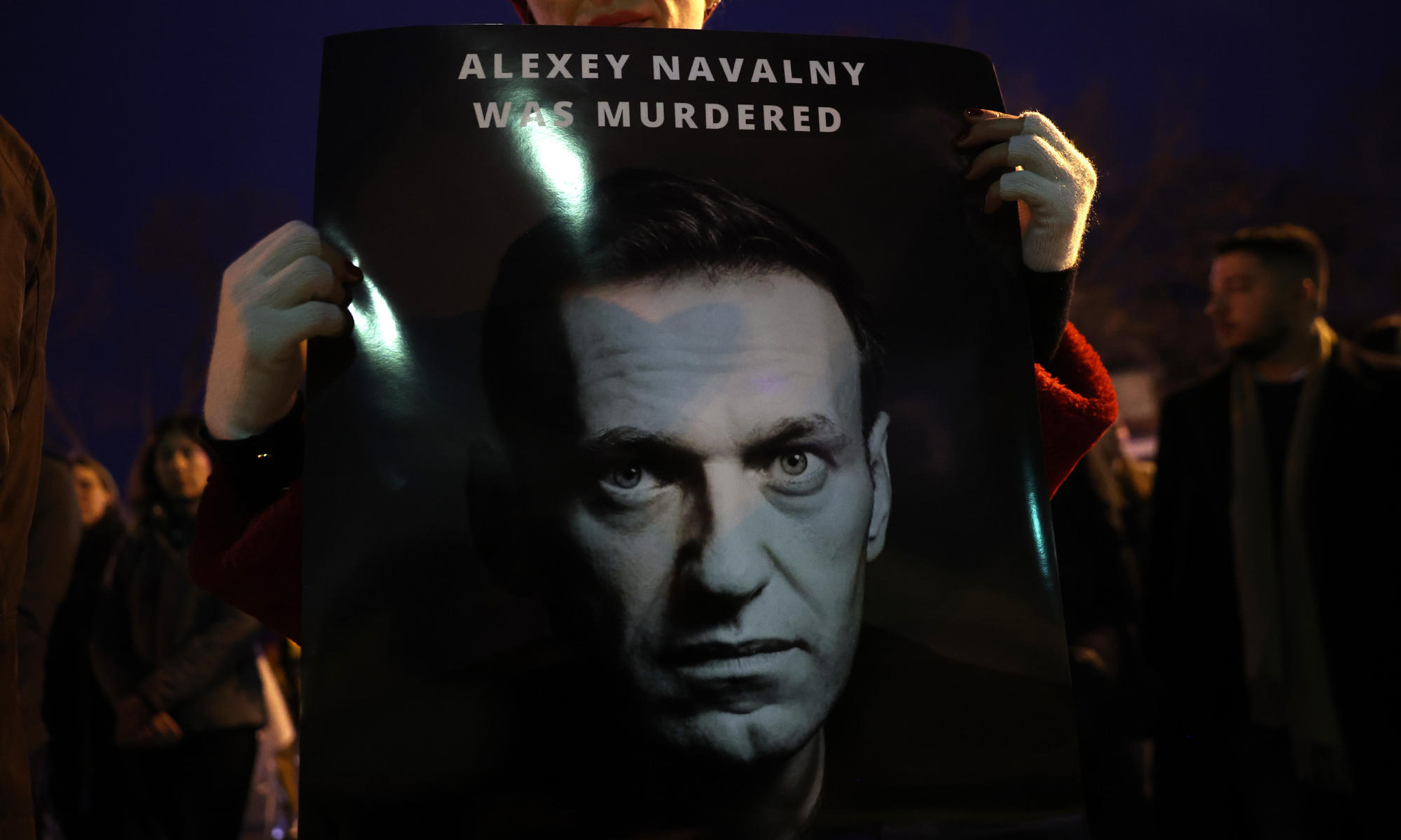 Supporters hold up a poster featuring the face of Alexei Navalny and the words "Alexei Navalny was murdered" during a nighttime vigil.