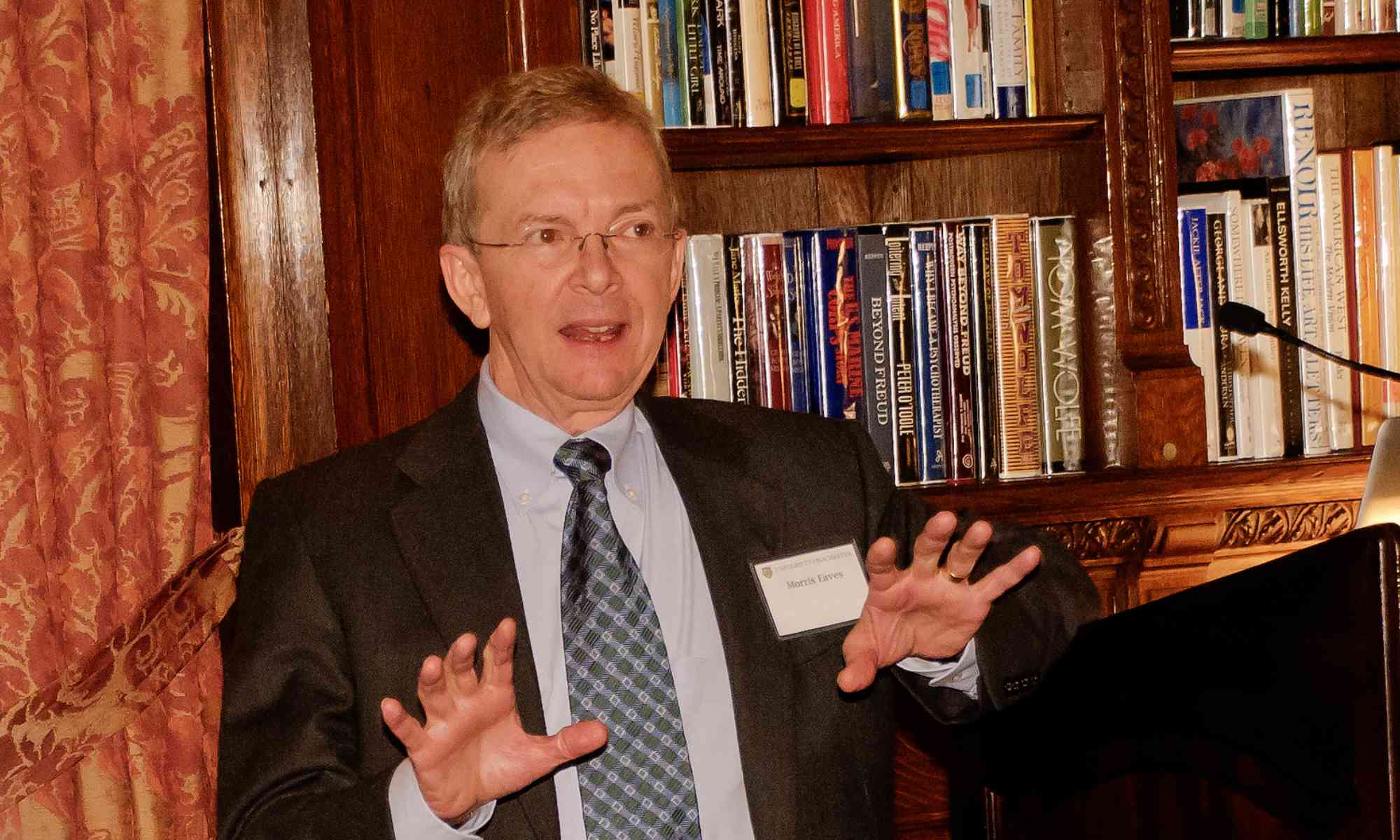 Morris Eaves speaking and gesturing at a lectern in a room reminiscent of a library during a ceremony.