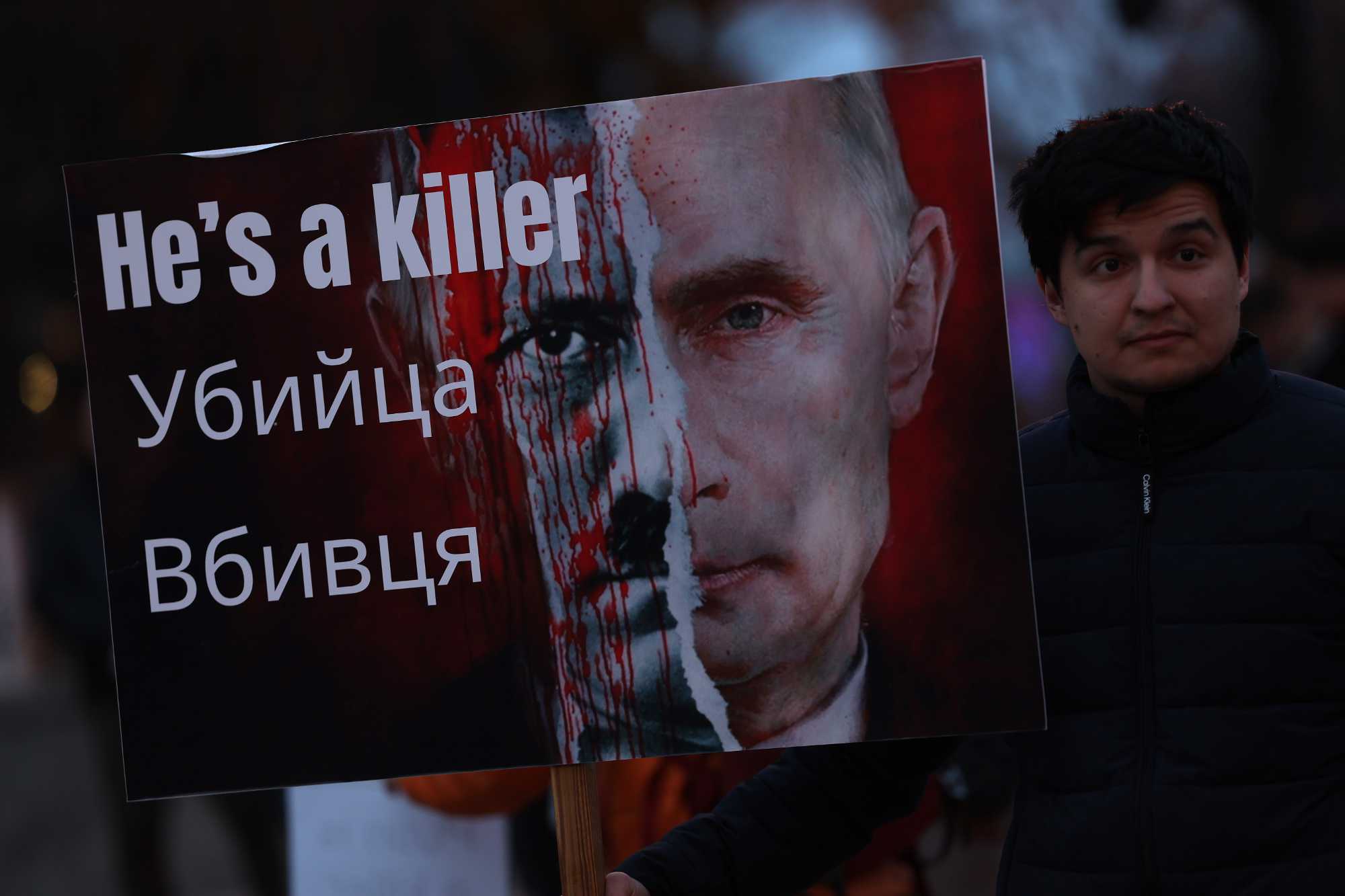 Close up of a person holding a protest sign with Vladimir Putin's face on it that says "He's a killer" in English and Russian to illustrate opposition movements.