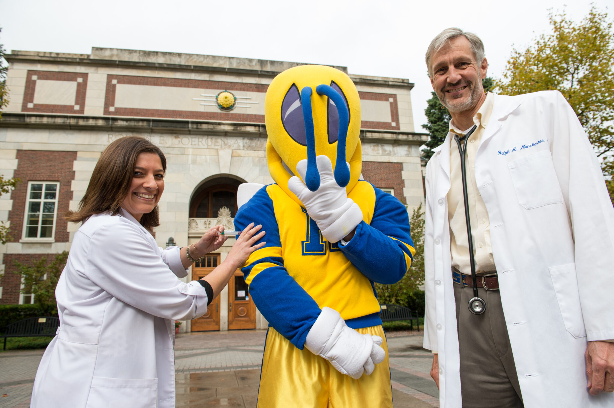 Two health care workers in white coats administered a flu shot to University of Rochester mascot Rocky.