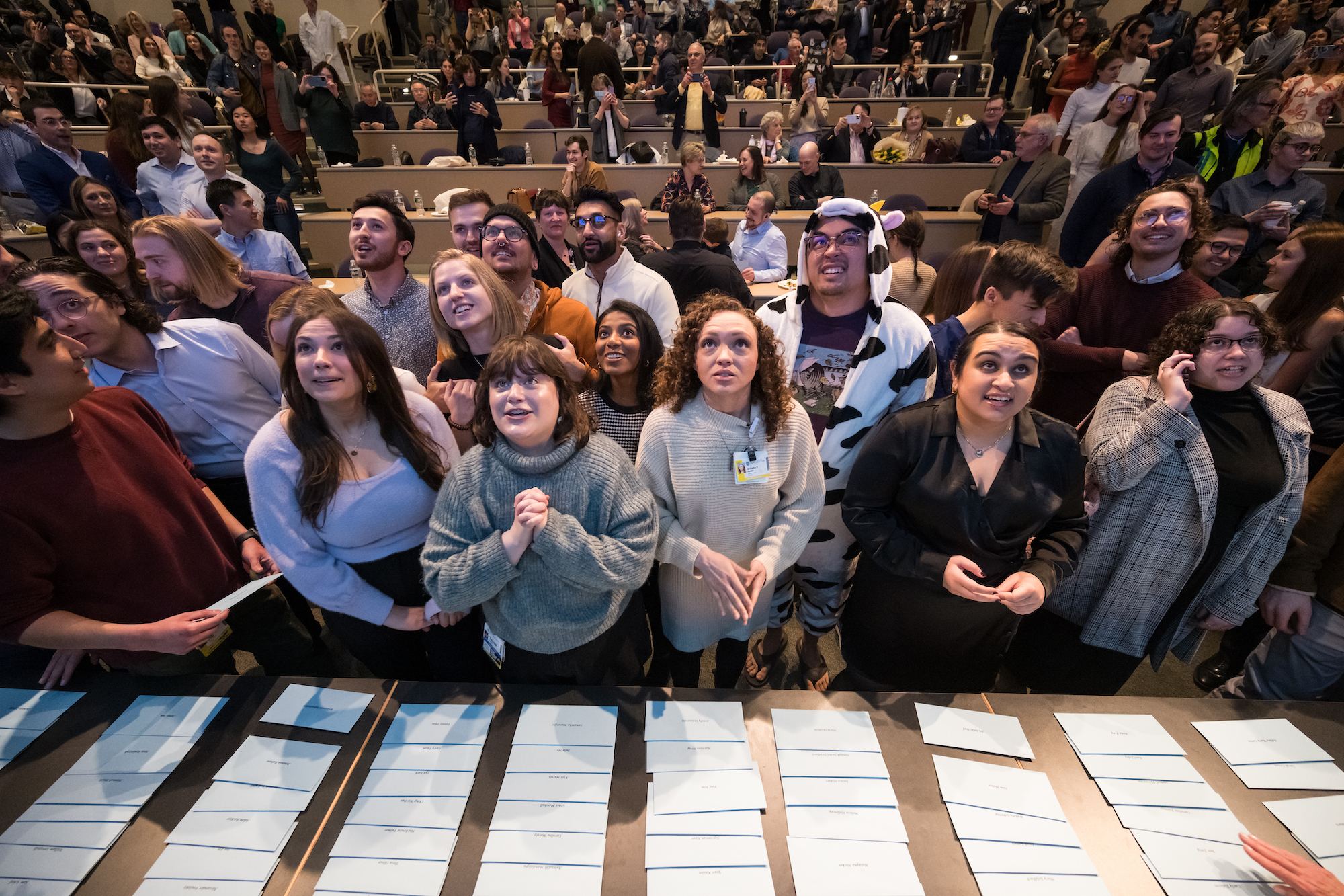 A roomful of students stands anxiously behind a table with white envelopes