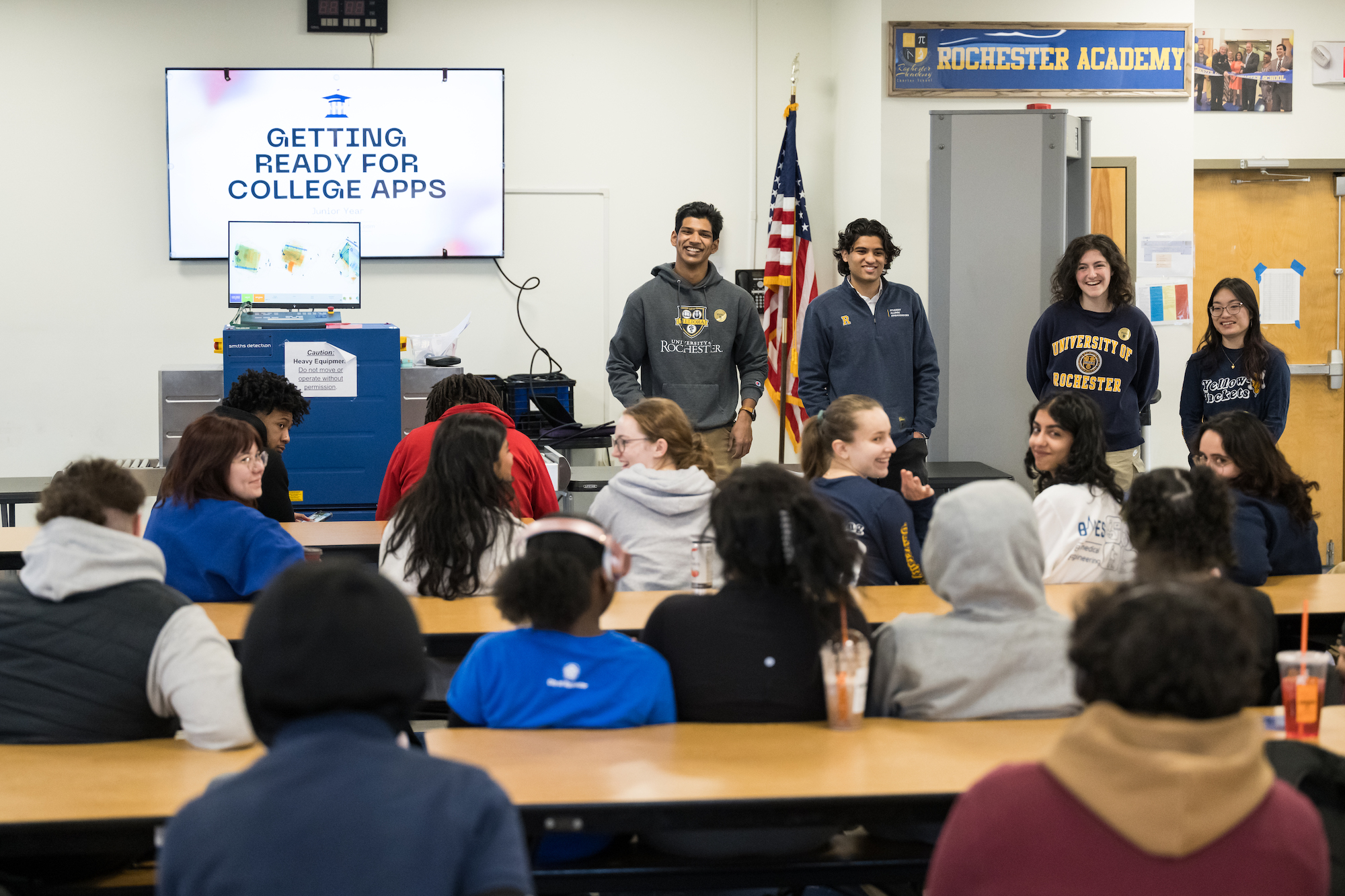 Four college students stand at the front of a high school class next to a white board that reads "Getting Ready for College Apps"