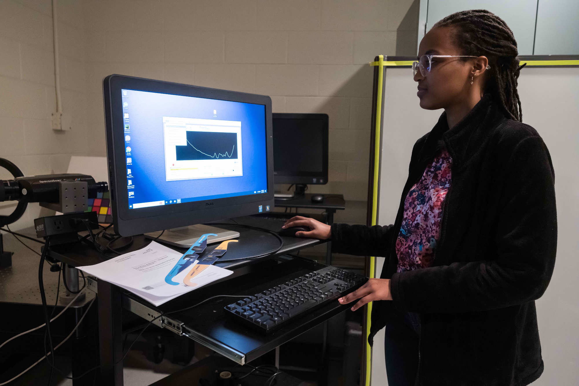 Graduate student stands at a computer analyzing data about solar eclipse sunglasses that are placed nearby.