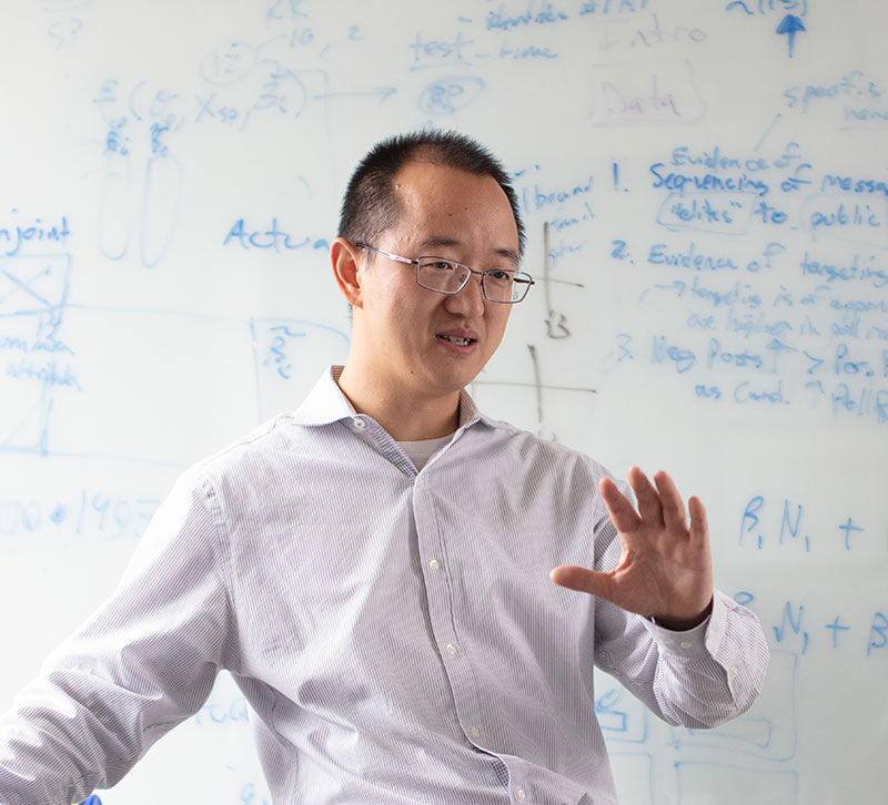 Huaxia Rui stands and gestures in front of a white board filled with equations and numbers written in blue marker.