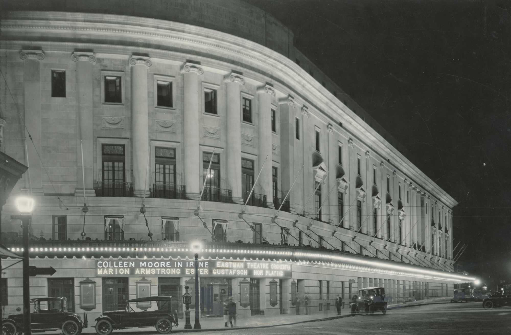 Vintage black-and-white image of Eastman Theatre at night with 1920s vehicles outside of it and the marquee lit up with the names of the performers.