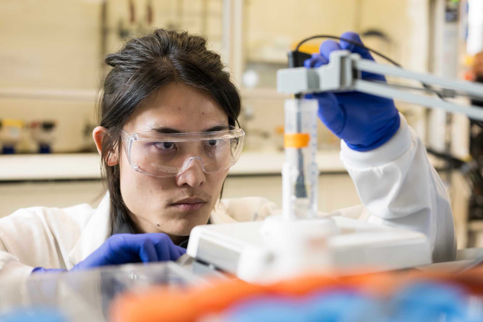 A scientist wearing lab googles and gloves examines a beaker of water containing PFAS chemicals.