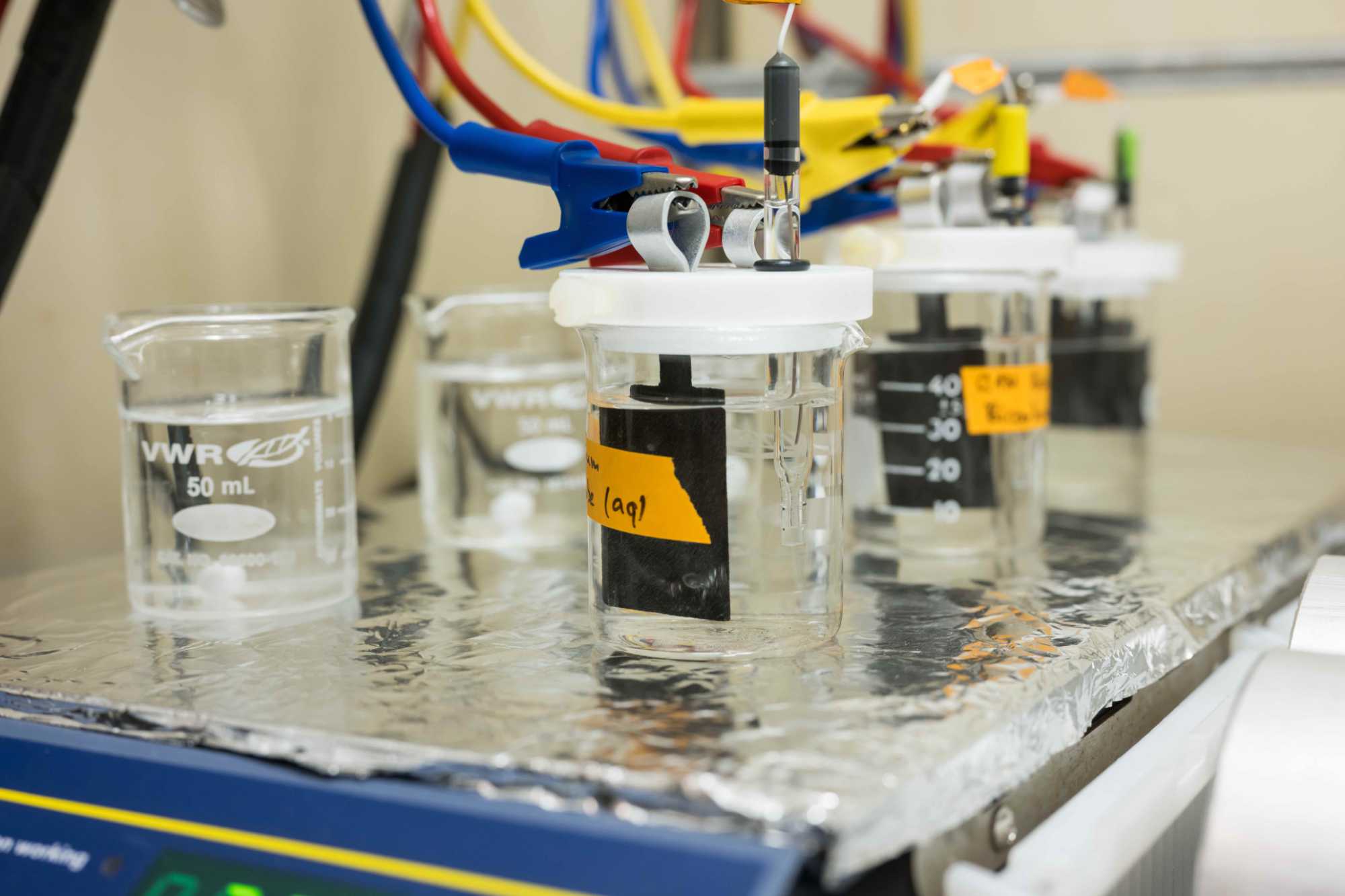 PFAS chemical remediation depicted with five beakers filled with water and hydrophilic carbon paper with blue, red, and yellow wires and jumper cable-style clamps affixed to them, all atop a scale covered with aluminum foil.