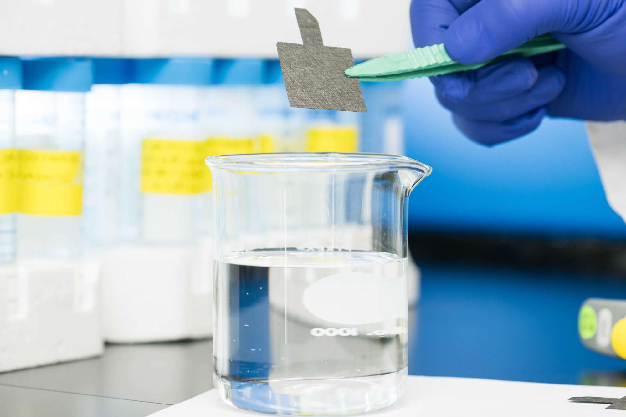 A gloved hands uses tweezers to hold carbon paper above a glass beaker filled with water to illustrate a new technique to remediate PFAS chemicals.